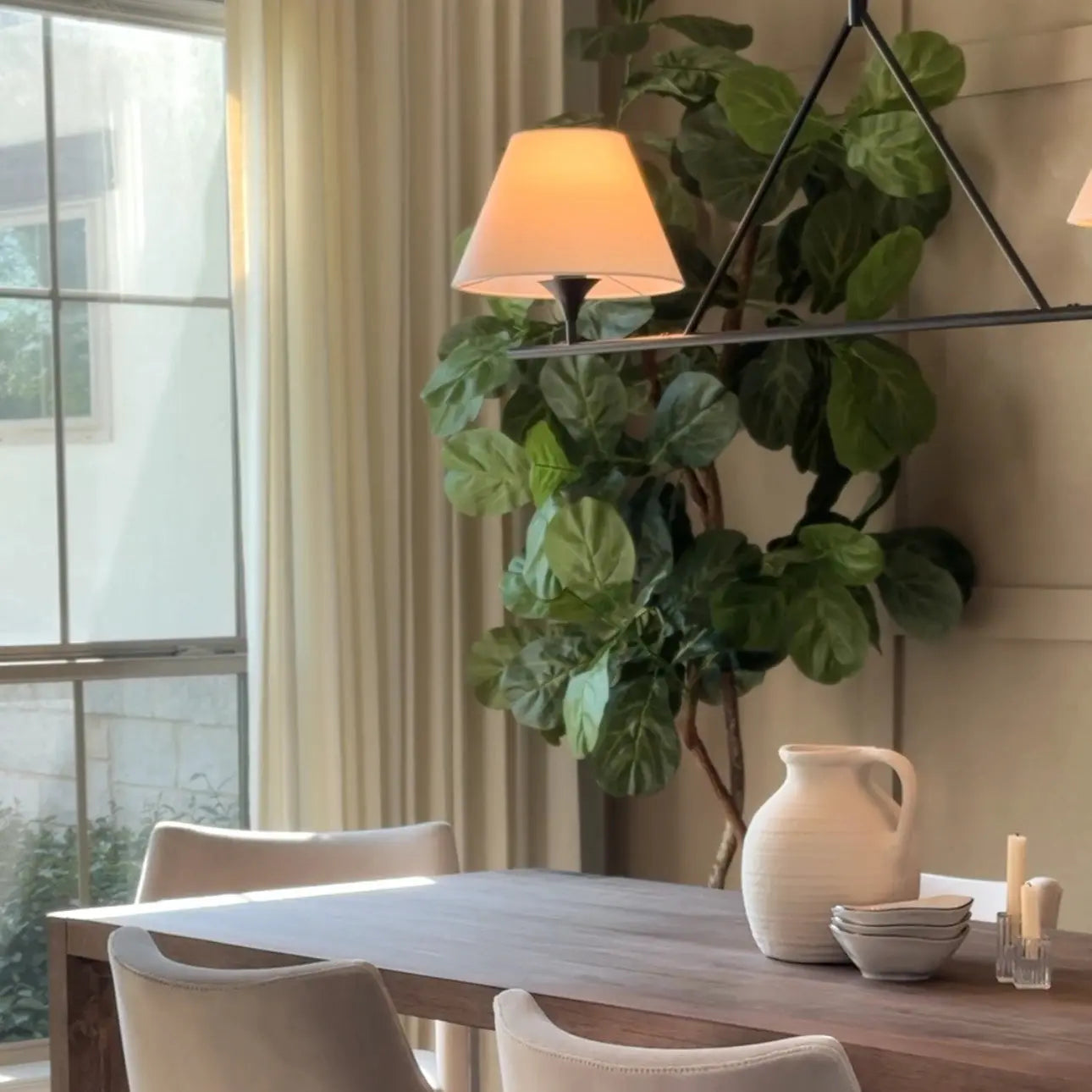 A bright photograph of a modern dining room featuring a prominent fiddle leaf fig tree, a linear chandelier with two illuminated lampshades, and a textured white ceramic pitcher with small bowls on a dark wooden table near a sunlit window