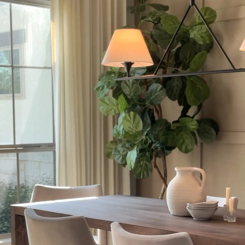 A bright photograph of a modern dining room featuring a prominent fiddle leaf fig tree, a linear chandelier with two illuminated lampshades, and a textured white ceramic pitcher with small bowls on a dark wooden table near a sunlit window