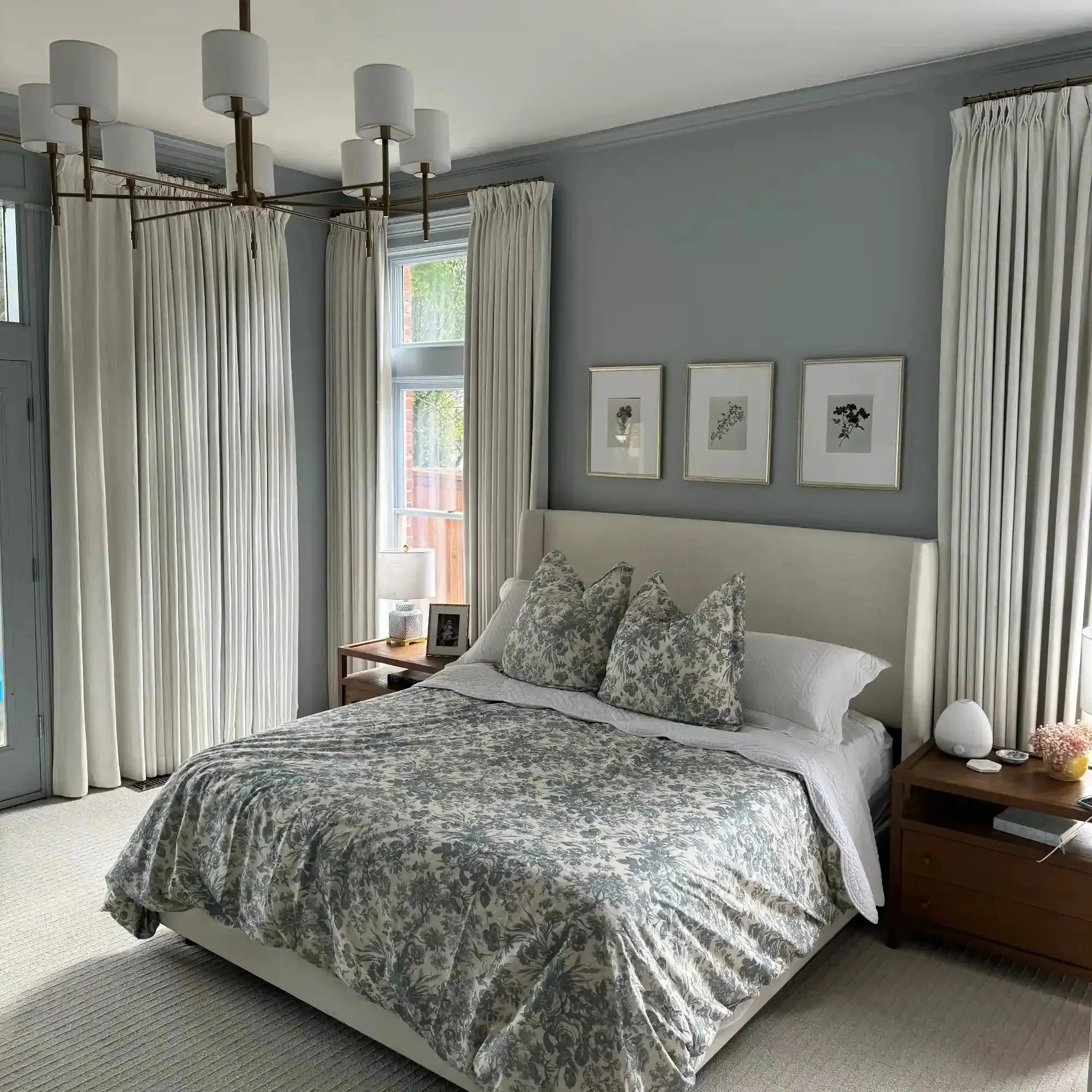 A modern bedroom showcases a white upholstered bed with a gray floral duvet and throw pillows, a striking bronze sputnik-style chandelier, and three framed botanical prints above the headboard on light gray walls