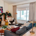 A cozy, festively decorated living room with a brick fireplace draped in Christmas garland, a red YOU neon sign, a dark grey sectional sofa, a brass and woven floor lamp, and a large window revealing a snowy outdoor patio