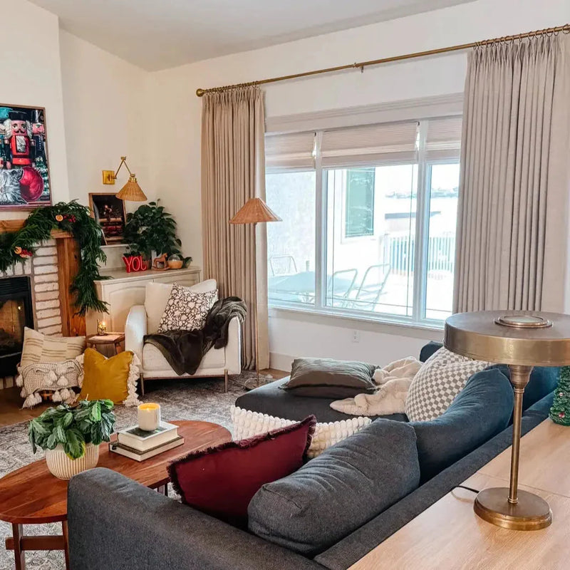 A cozy, festively decorated living room with a brick fireplace draped in Christmas garland, a red YOU neon sign, a dark grey sectional sofa, a brass and woven floor lamp, and a large window revealing a snowy outdoor patio