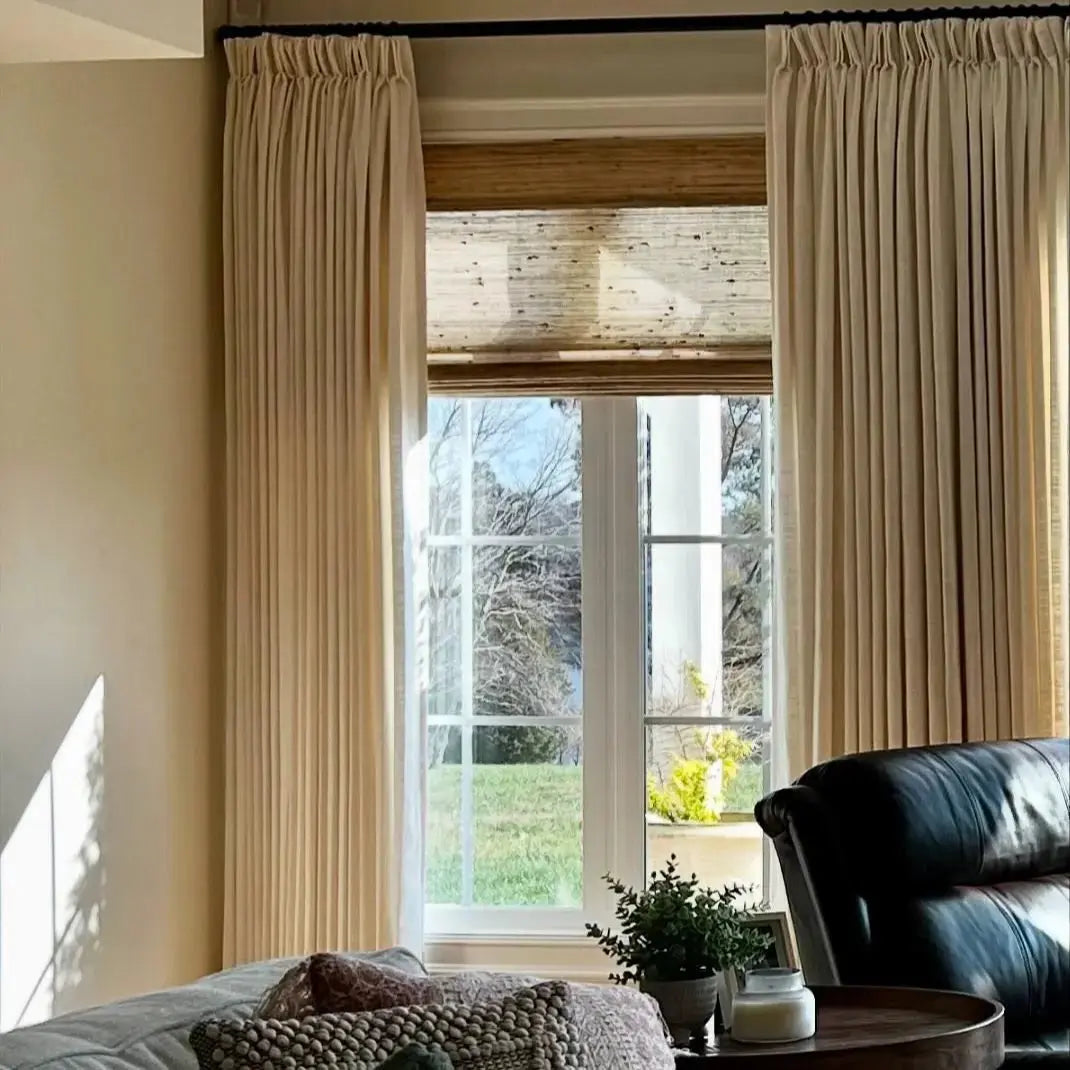 Interior view of a living room window with beige pleated drapes and a natural woven wood blind, revealing a sunny green lawn, bare trees, and a distant white architectural column, with a dark leather armchair and patterned pillows in the foreground
