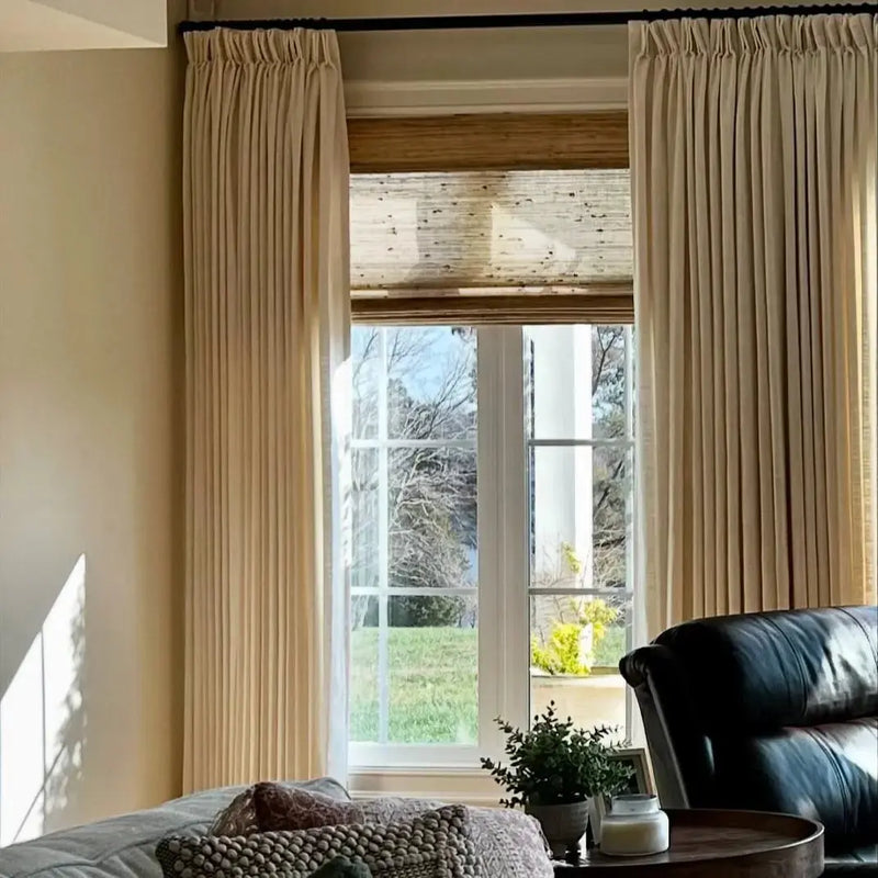 Interior view of a living room window with beige pleated drapes and a natural woven wood blind, revealing a sunny green lawn, bare trees, and a distant white architectural column, with a dark leather armchair and patterned pillows in the foreground