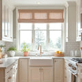 Photograph of a bright U-shaped kitchen featuring white shaker cabinets, a farmhouse sink under a large window with a tan Roman shade, white subway tile backsplash, and brushed gold hardware