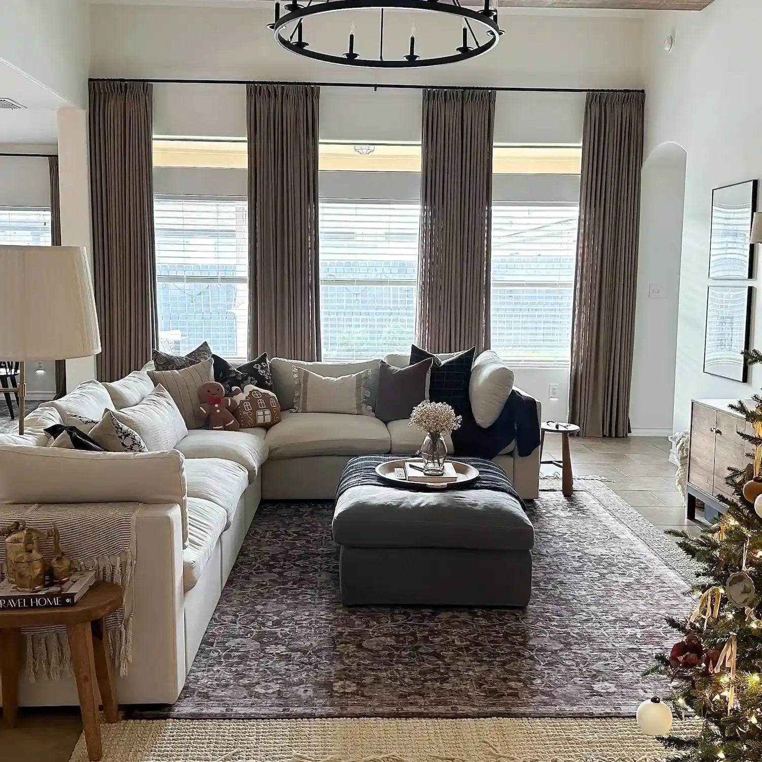 Interior photograph of a modern living room centered around a large white sectional sofa and a striking black circular chandelier with tall windows framed by brown drapes a small lit Christmas tree and charming gingerbread house pillows adding festive touches to the patterned rug