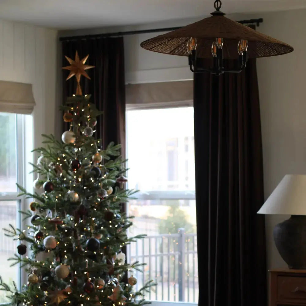 Indoor scene with a decorated Christmas tree topped with a golden star and adorned with white and gold ornaments, beside a distinct woven straw chandelier and rich brown velvet curtains