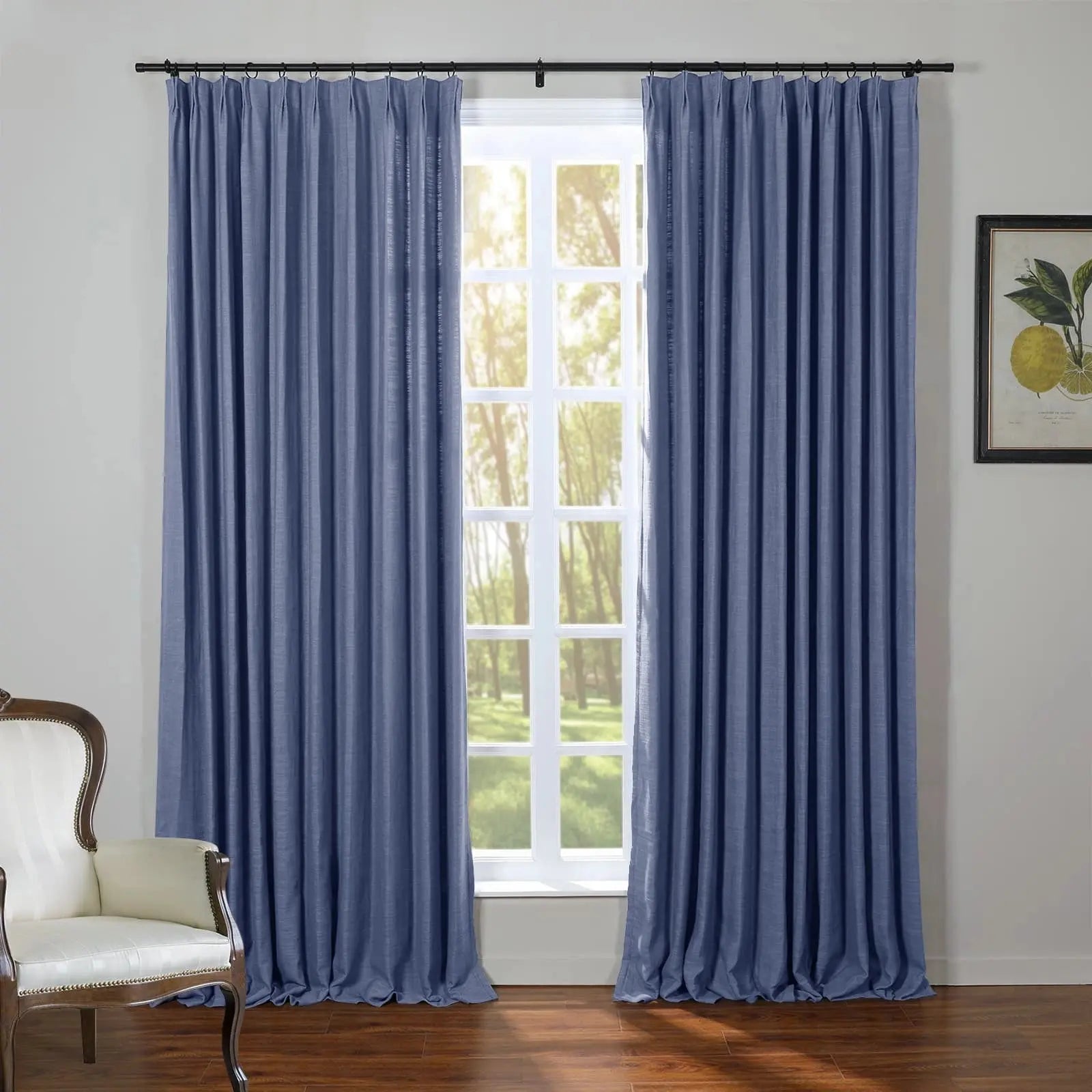 A room features floor-length indigo blue textured pinch-pleat curtains on a black rod, framing a white paned window with sunlit green trees outside, alongside a dark hardwood floor, a white antique armchair, and a framed botanical lemon print
