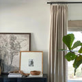 An artfully styled home interior with a dark cabinet displaying a monochromatic landscape print, a framed child's photo, stacked books, a wooden chain, and a woven basket, positioned next to a window with neutral beige drapery and a vibrant fiddle leaf fig plant