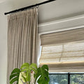 A bright interior window scene featuring beige textured drapes on a black rod, a woven Roman blind, and a vibrant variegated Monstera plant with large split green leaves