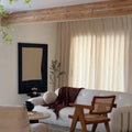 A stylish minimalist living room featuring a white boucl¨¦ sofa with a dark brown throw, a large abstract black and beige artwork, and a prominent cane and wood chair under a wooden beam ceiling
