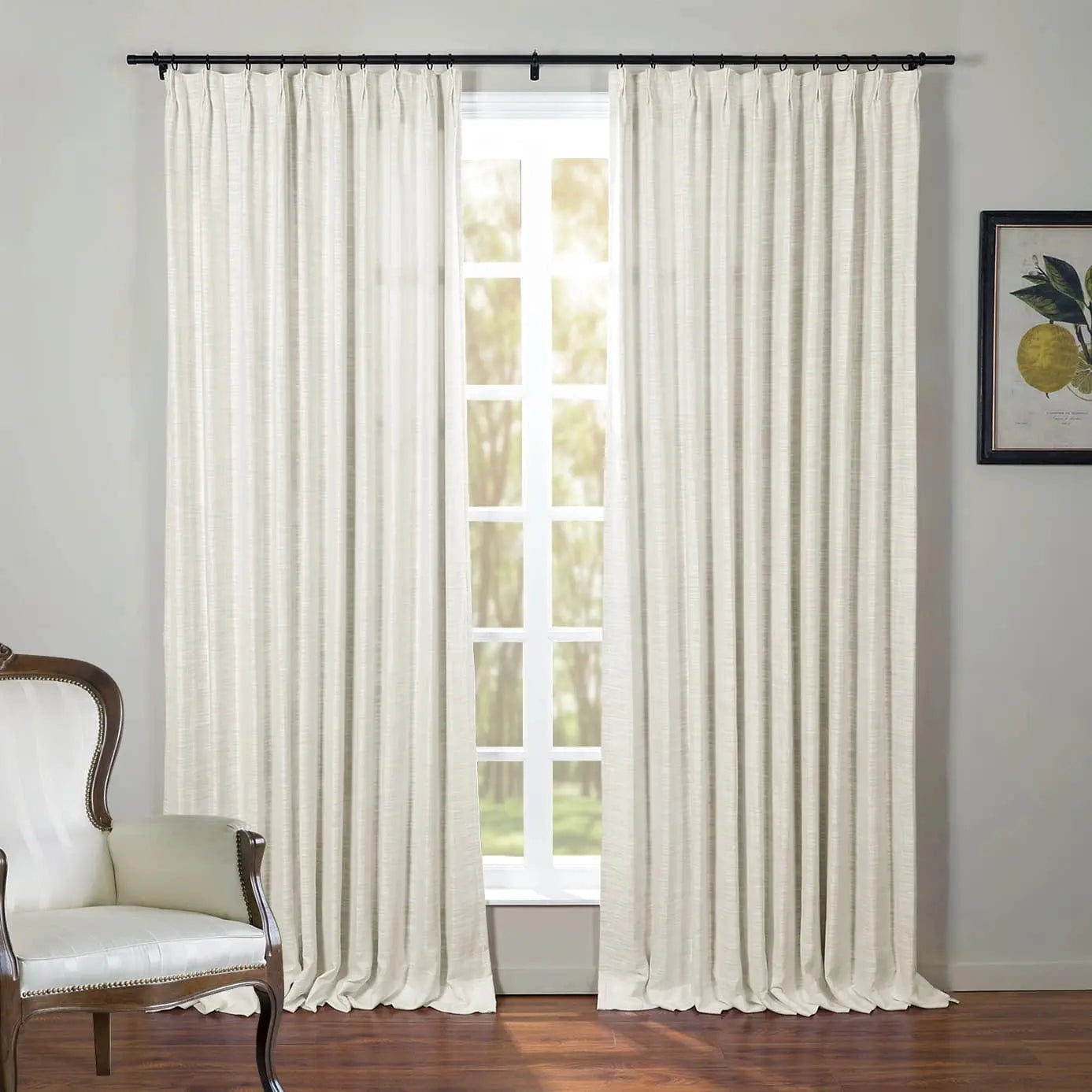 Cream linen-look pinch-pleat curtains frame a sunlit window with trees outside, an elegant white upholstered armchair with dark wood and nailhead trim sits to the left, and a framed lemon botanical print adorns the right wall