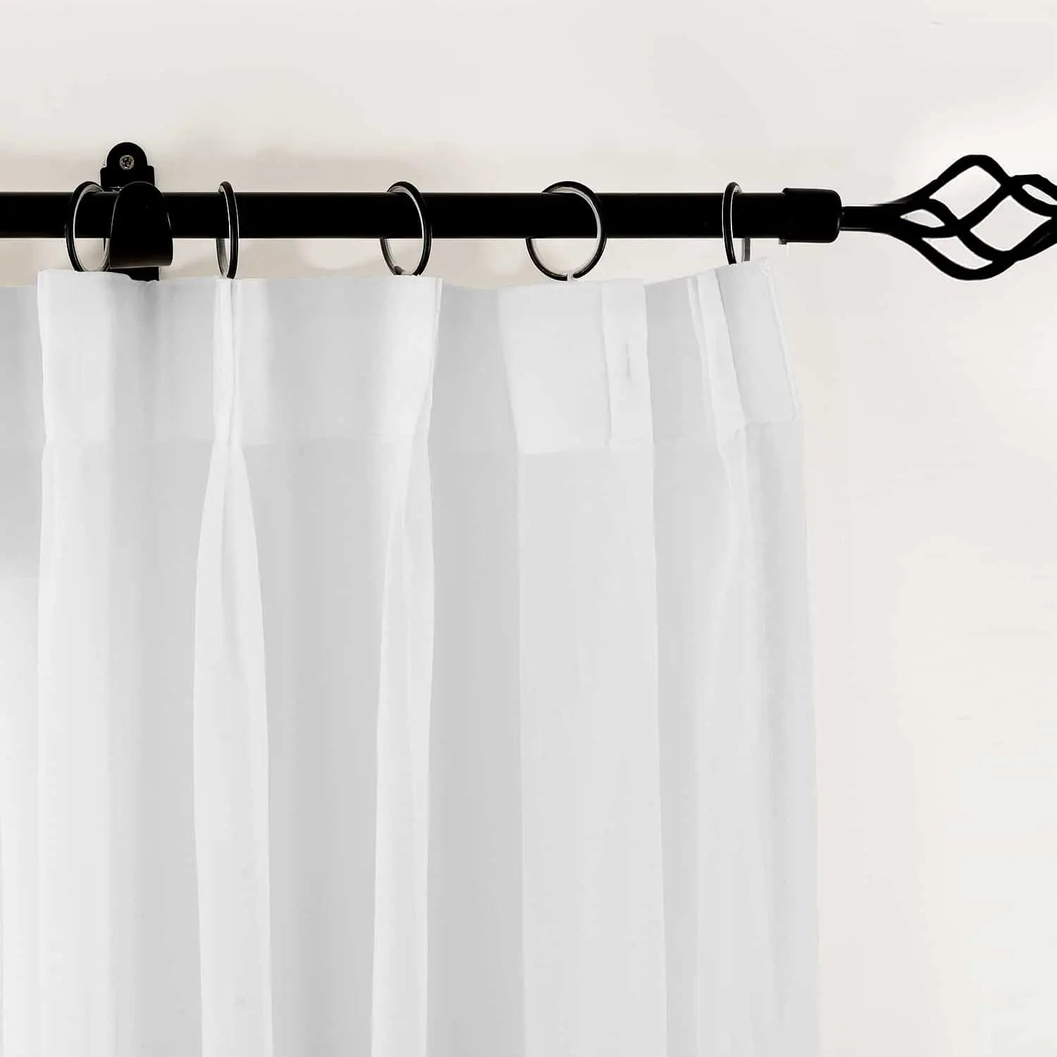 Close-up of a white sheer curtain panel hanging from a black wrought iron curtain rod with a decorative twisted cage finial and metal rings against a plain light wall