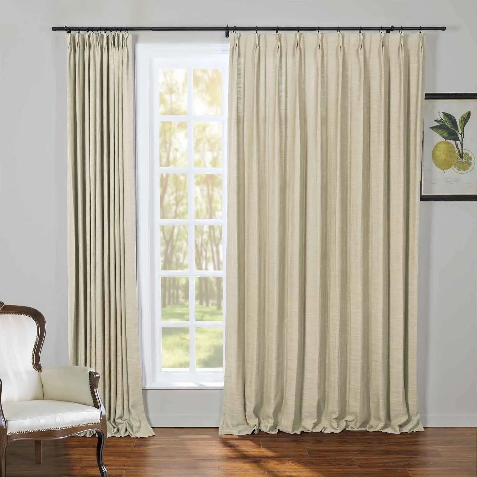 Beige textured pinch-pleat curtains on a black rod in a bright room with a white grid window overlooking a sunny forest, a cream upholstered armchair with dark wood trim, a framed lemon botanical print, and dark hardwood floors
