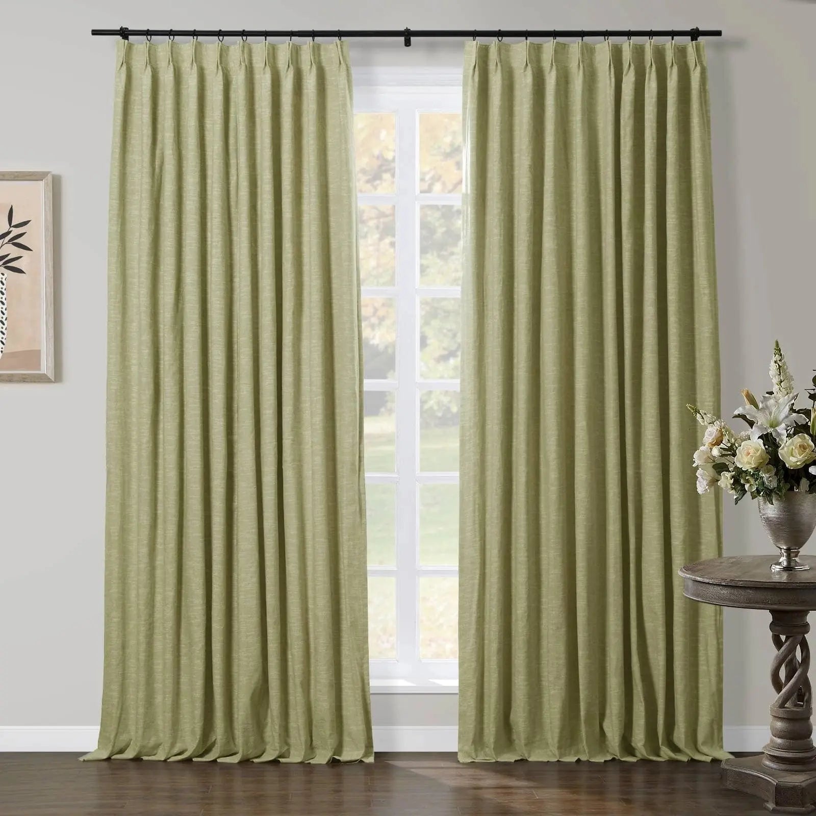 Light green linen-textured curtains hung on a black rod framing a window in a room, with a twisted wooden accent table holding a silver vase of white lilies and cream roses