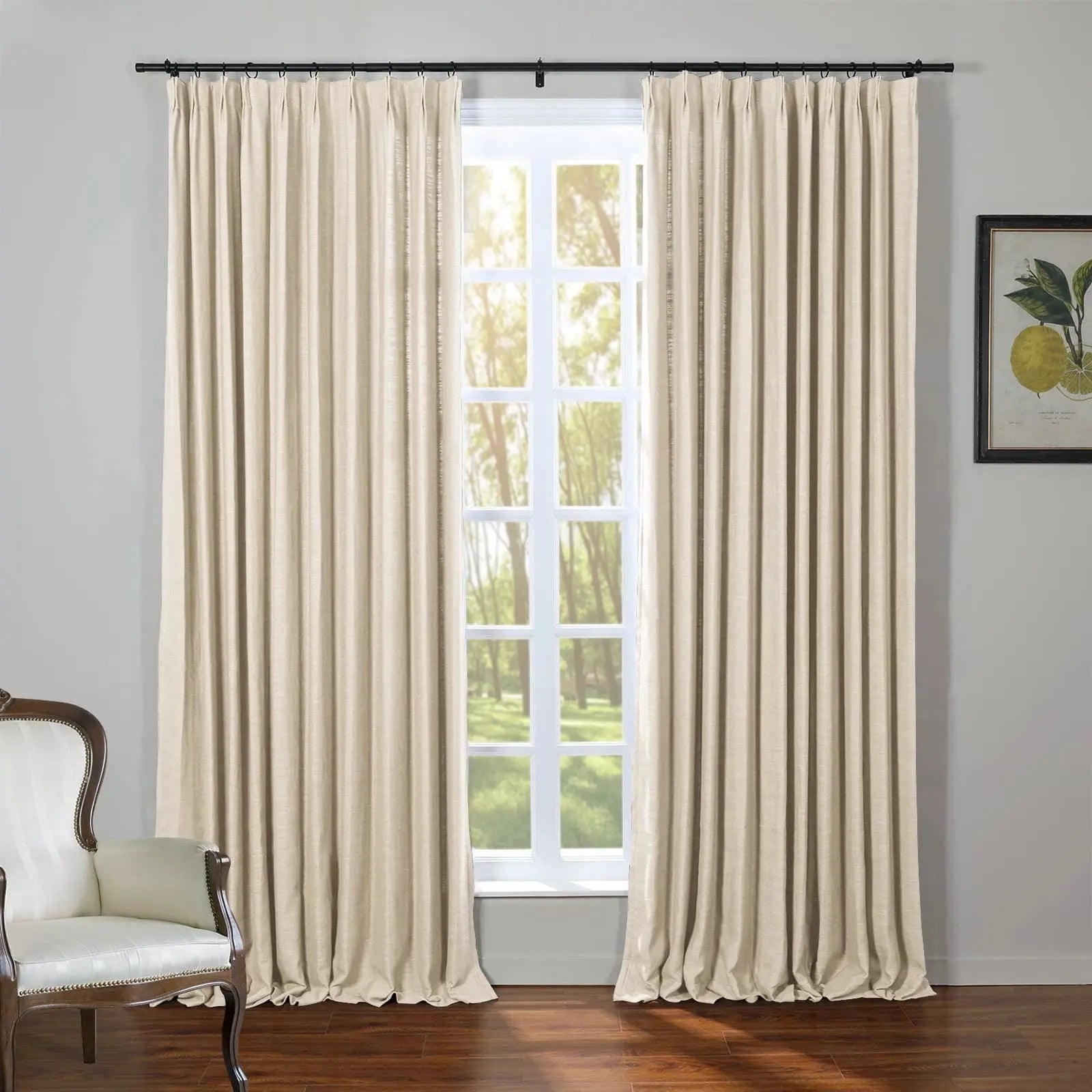 Photograph of an interior room with elegant beige pinch-pleat linen curtains framing a bright window overlooking lush green trees, complemented by a classic white armchair with dark wood accents and a botanical lemon print on the gray wall