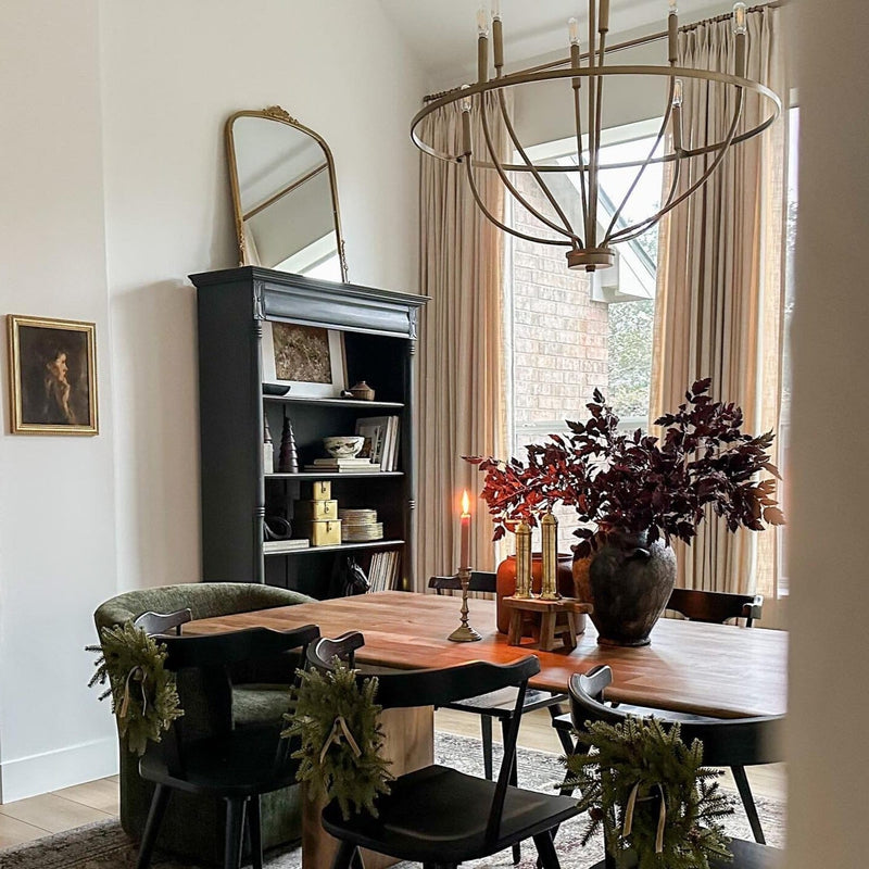 Photograph of a festive dining room featuring a long wooden table with black chairs adorned with evergreen wreaths, a large vase of deep red foliage, a lit candle, and an ornate brass chandelier hanging above, with a dark bookshelf and gold-framed mirror against the wall