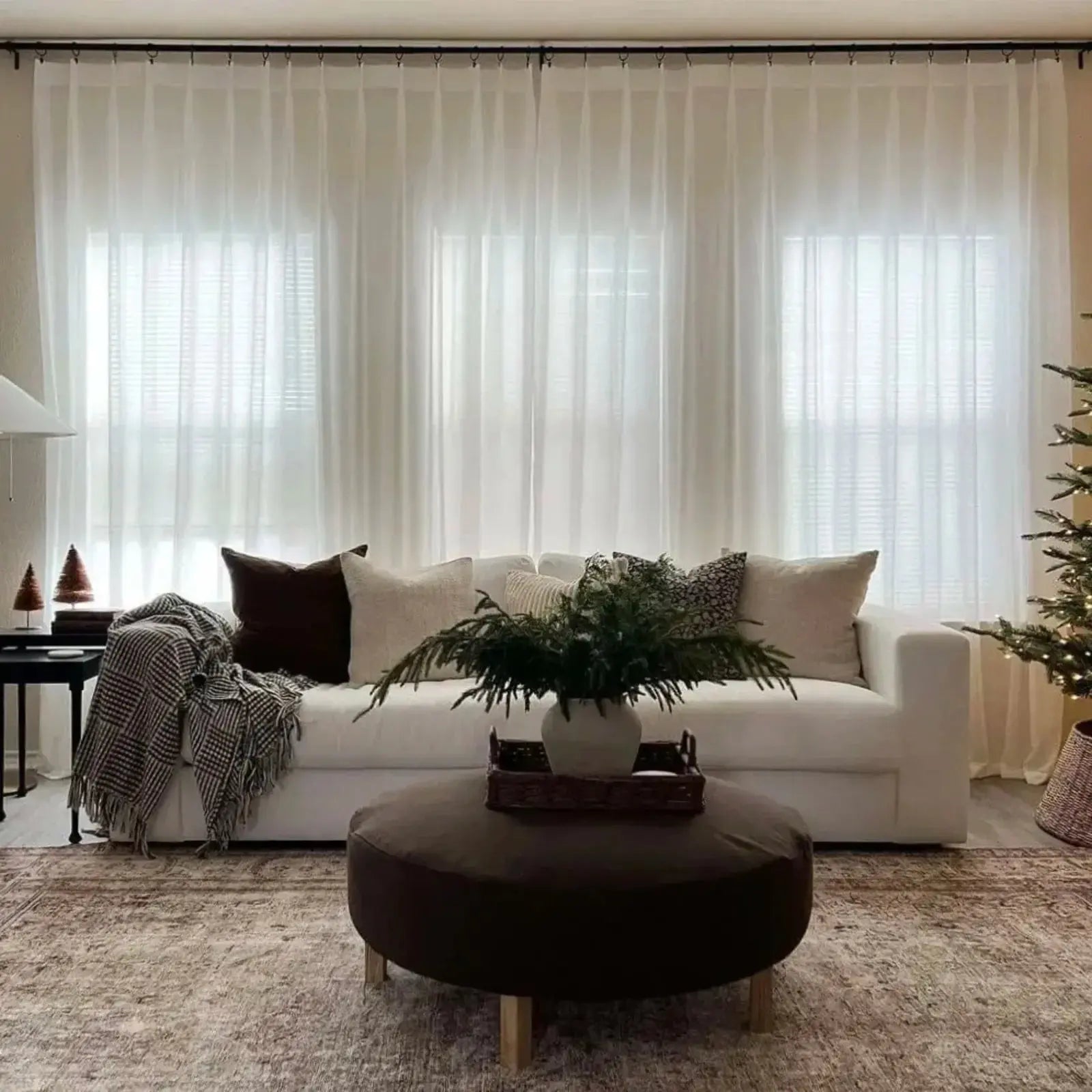 A cozy modern living room with a white sofa adorned with chocolate brown and cream pillows, a dark brown round ottoman holding a vase of evergreen branches on a patterned rug, sheer white curtains, and a small illuminated Christmas tree