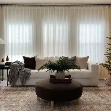 A cozy modern living room with a white sofa adorned with chocolate brown and cream pillows, a dark brown round ottoman holding a vase of evergreen branches on a patterned rug, sheer white curtains, and a small illuminated Christmas tree