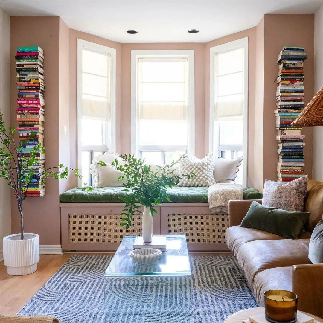 A cozy blush-pink living room with a bay window featuring a green velvet cushioned seat with woven cane details, flanked by tall vertical book stacks on the walls, and a tan leather sofa