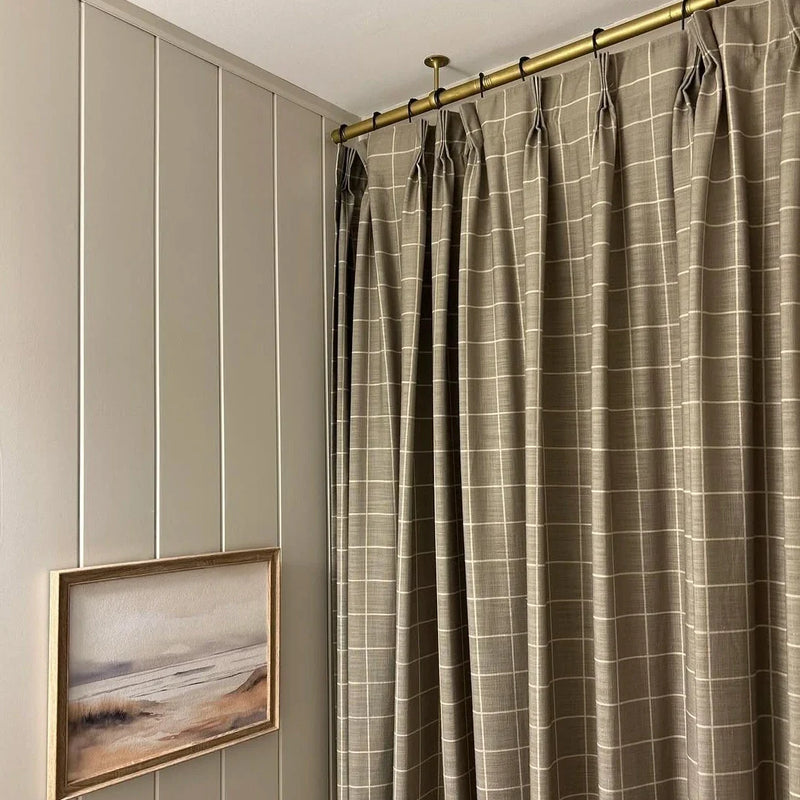 Photograph of an interior room showcasing taupe windowpane plaid curtains with pinch pleats on a brass rod beside light greige board and batten wall paneling displaying a framed muted watercolor landscape painting