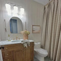 A neutral-toned modern farmhouse bathroom featuring white shiplap walls, a light wood vanity with a marble countertop and brass fixtures, an arched mirror, a peach vase holding dried hydrangeas, and a tan grid-patterned shower curtain