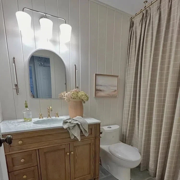 A neutral-toned modern farmhouse bathroom featuring white shiplap walls, a light wood vanity with a marble countertop and brass fixtures, an arched mirror, a peach vase holding dried hydrangeas, and a tan grid-patterned shower curtain