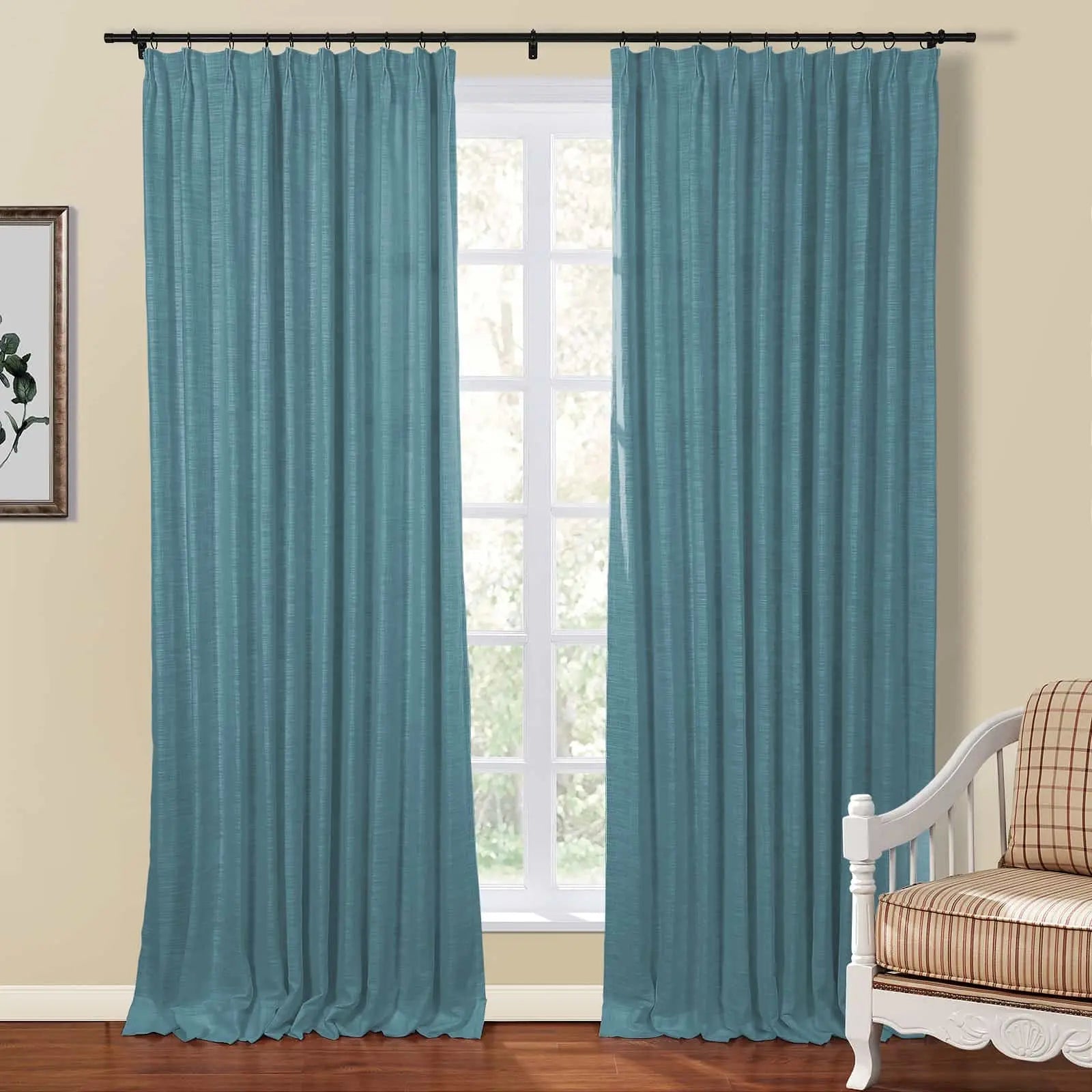 A pair of teal textured pinch pleat curtains on a black rod flank a white gridded window looking out to trees with a corner of a plaid armchair visible in an interior room