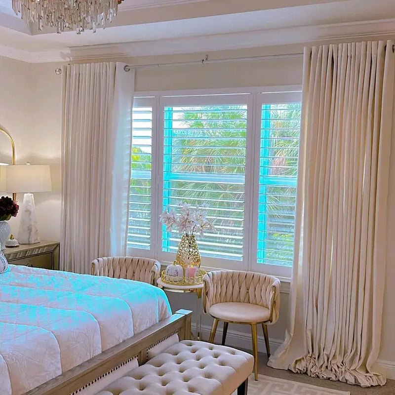 Interior photography depicting a luxurious contemporary bedroom with a crystal chandelier white plantation shutters opening to lush tropical palms and two beige ribbed velvet armchairs beside a gilded accent table