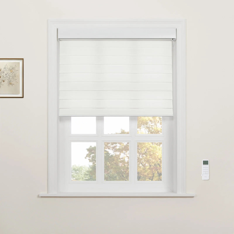 A white layered Roman Blind partially covers a multi-pane window in a neutral-toned room, featuring a wall-mounted remote control and a framed botanical drawing, with warm autumn trees visible outside