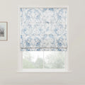 White Roman blind adorned with an intricate blue floral pattern partially covers a window, revealing blurred green foliage outdoors