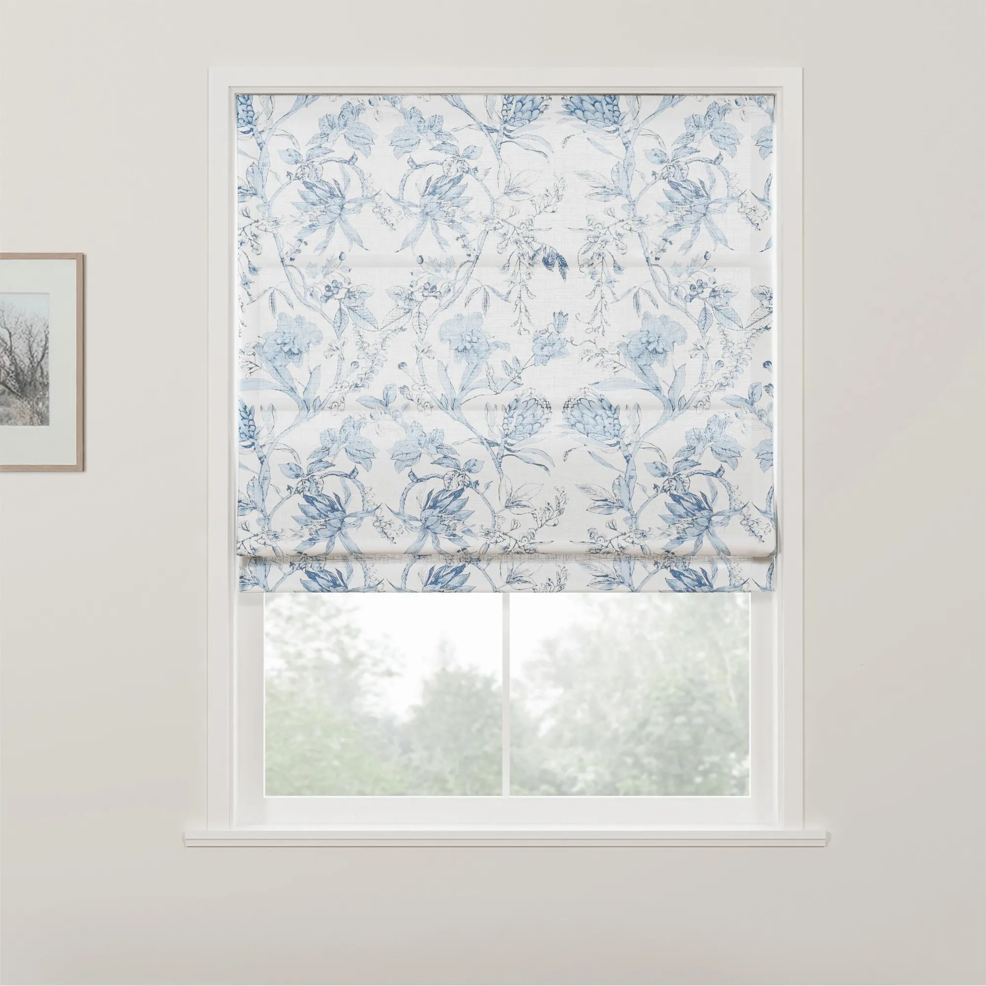 White Roman blind adorned with an intricate blue floral pattern partially covers a window, revealing blurred green foliage outdoors