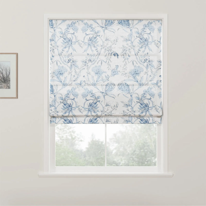 White Roman blind adorned with an intricate blue floral pattern partially covers a window, revealing blurred green foliage outdoors