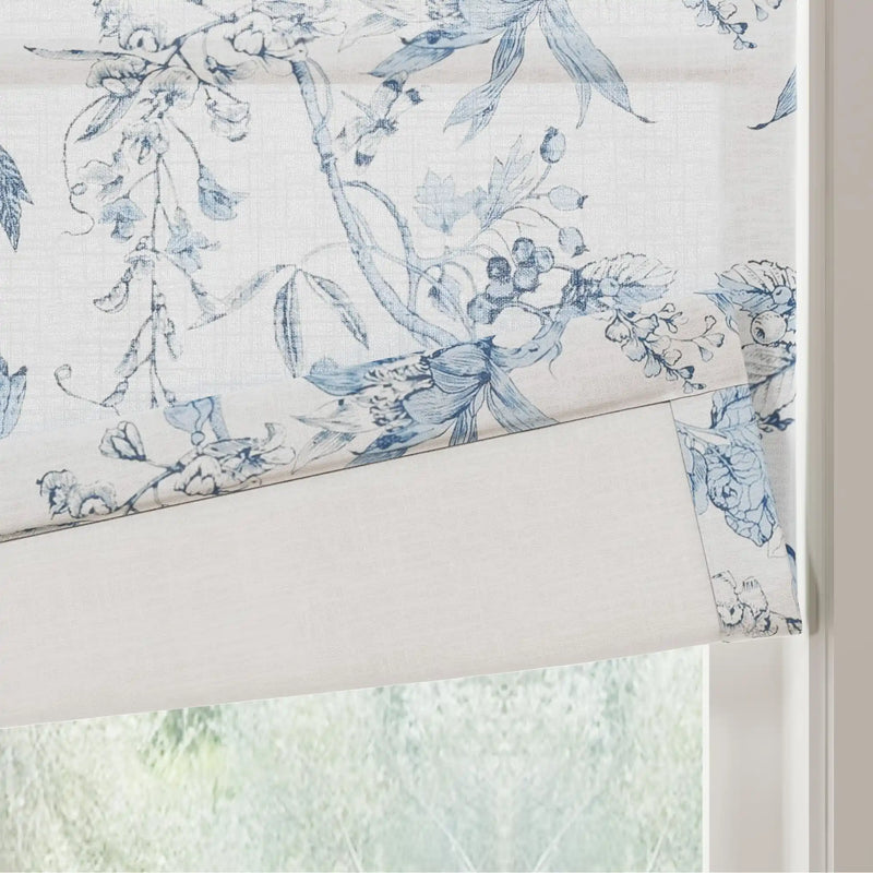 Photograph of a blue and white toile-style Roman blind featuring wisteria and berry patterns, partially lowered over a window with blurred green foliage outside