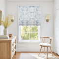 A bright interior room featuring a window with a blue floral Roman shade, a wooden dresser holding a vase of vibrant yellow flowers, and a light wood wishbone chair