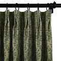 A close-up of a muted olive green curtain with a repeating dark grey leaf vine pattern hanging from a black metal rod with rings and distinct pleats