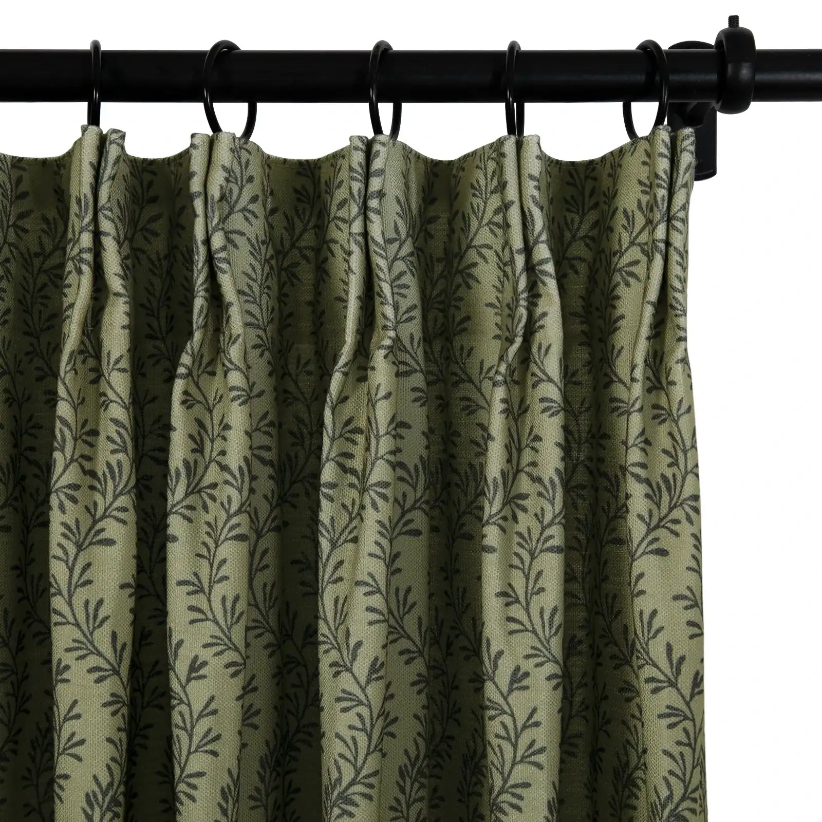 A close-up of a muted olive green curtain with a repeating dark grey leaf vine pattern hanging from a black metal rod with rings and distinct pleats