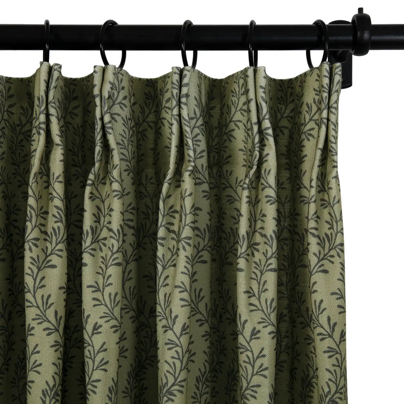 A close-up of a muted olive green curtain with a repeating dark grey leaf vine pattern hanging from a black metal rod with rings and distinct pleats