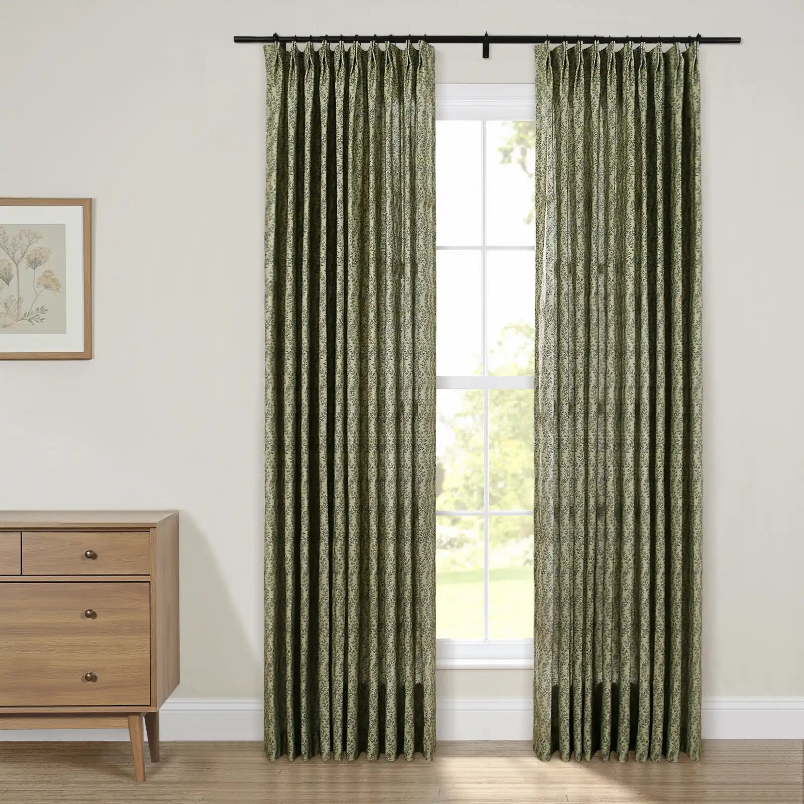 A pair of green botanical print pleated curtains framing a sunlit window in a neutral room interior featuring a light wooden dresser and framed botanical art