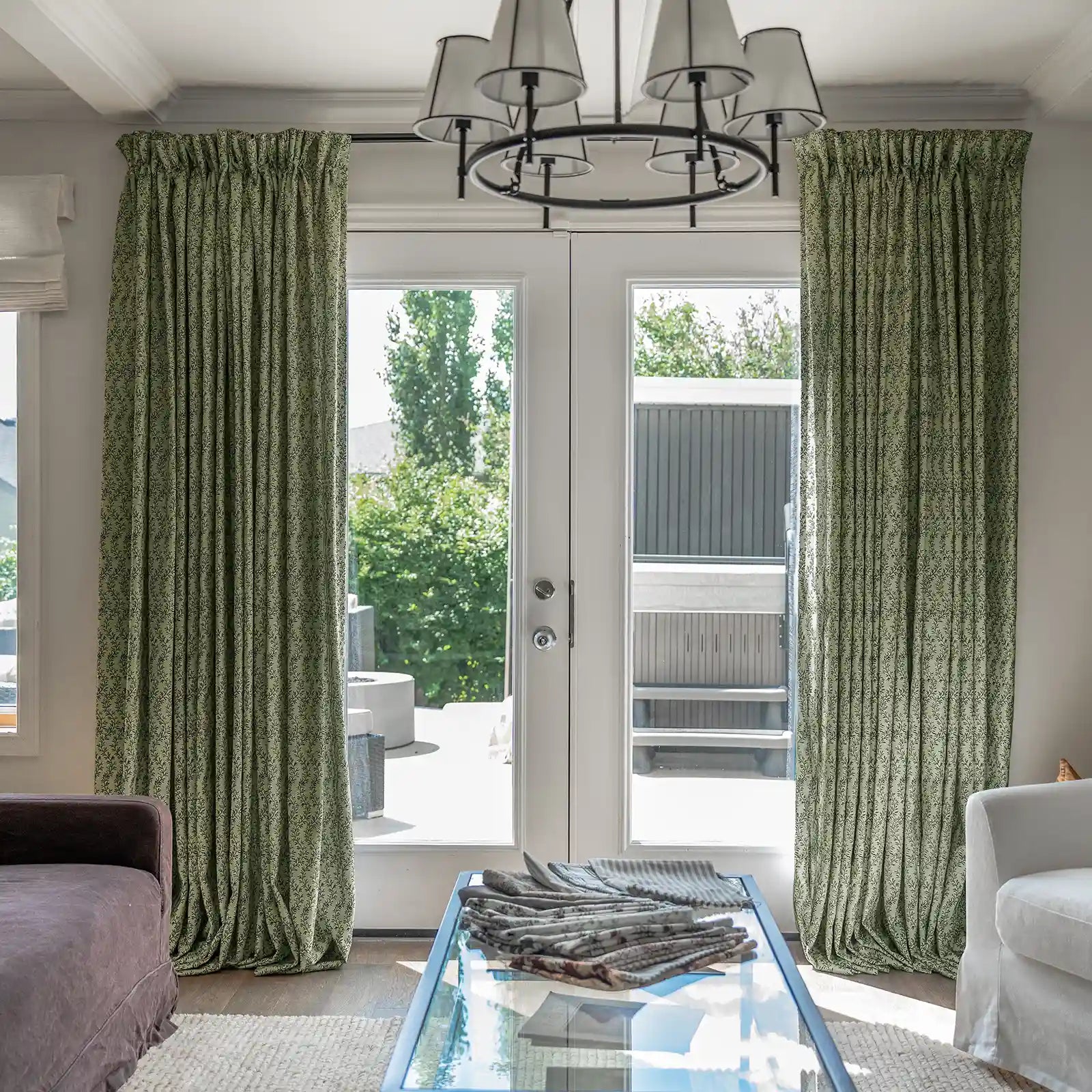 Interior living room features green patterned drapery, a black chandelier with white shades, and a glass coffee table with fabric swatches, looking out to a sunny patio with a hot tub