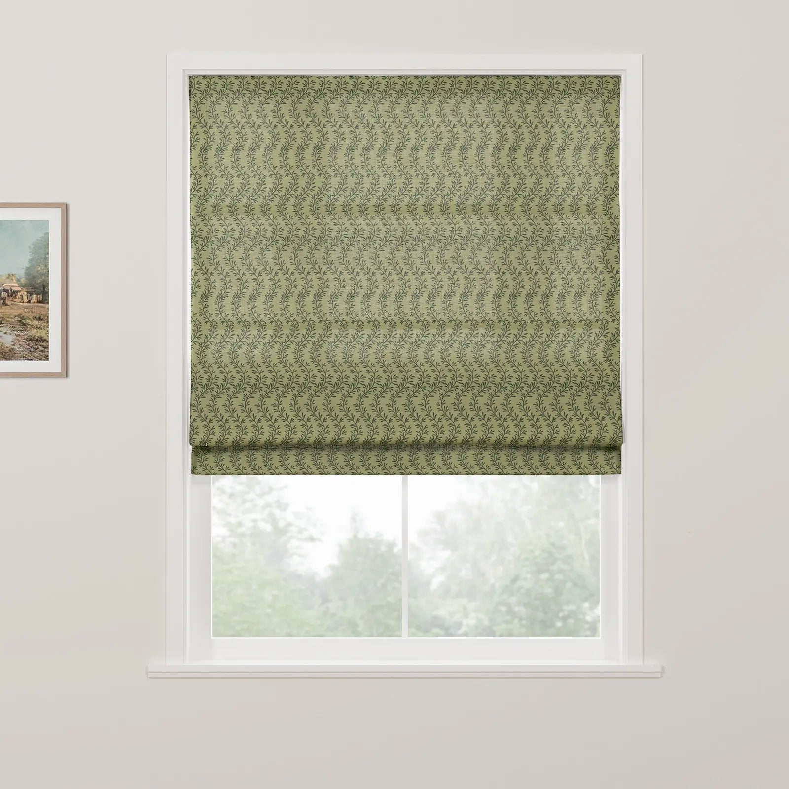 Roman blind featuring a muted green background with a delicate dark green leafy vine pattern partially drawn over a window offering a blurred view of trees, a framed rustic landscape painting hangs nearby