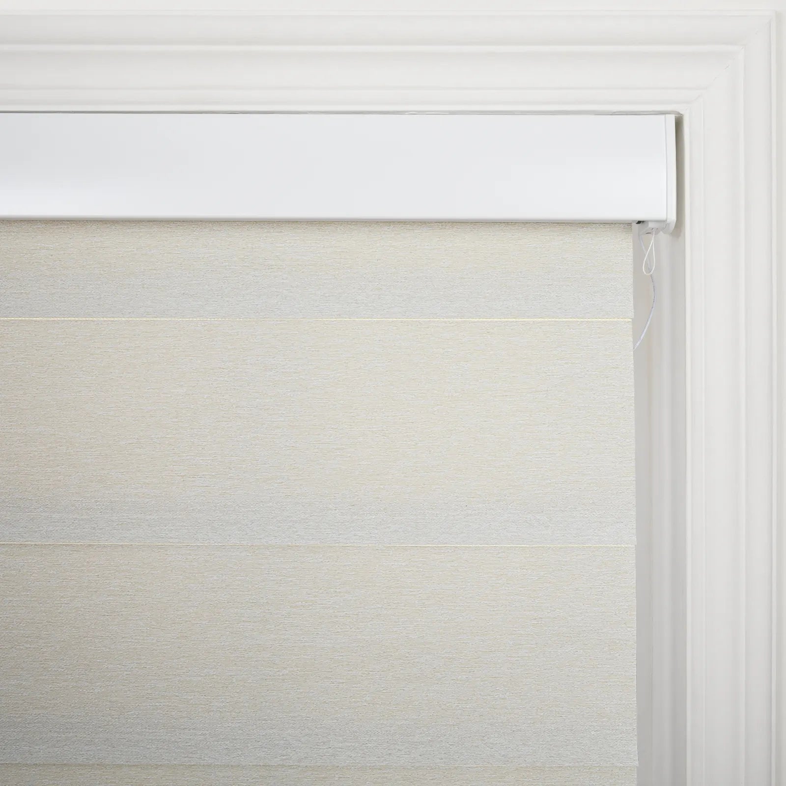 A close-up of a beige textured window Blind with subtle horizontal banding, a white headrail, and classic white decorative window trim, featuring a slender control cord visible on the right