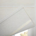 A textured light grey fabric window Blind partially unfurled, showcasing a subtle cream backing strip and a glimpse of blurred green-yellow foliage outside