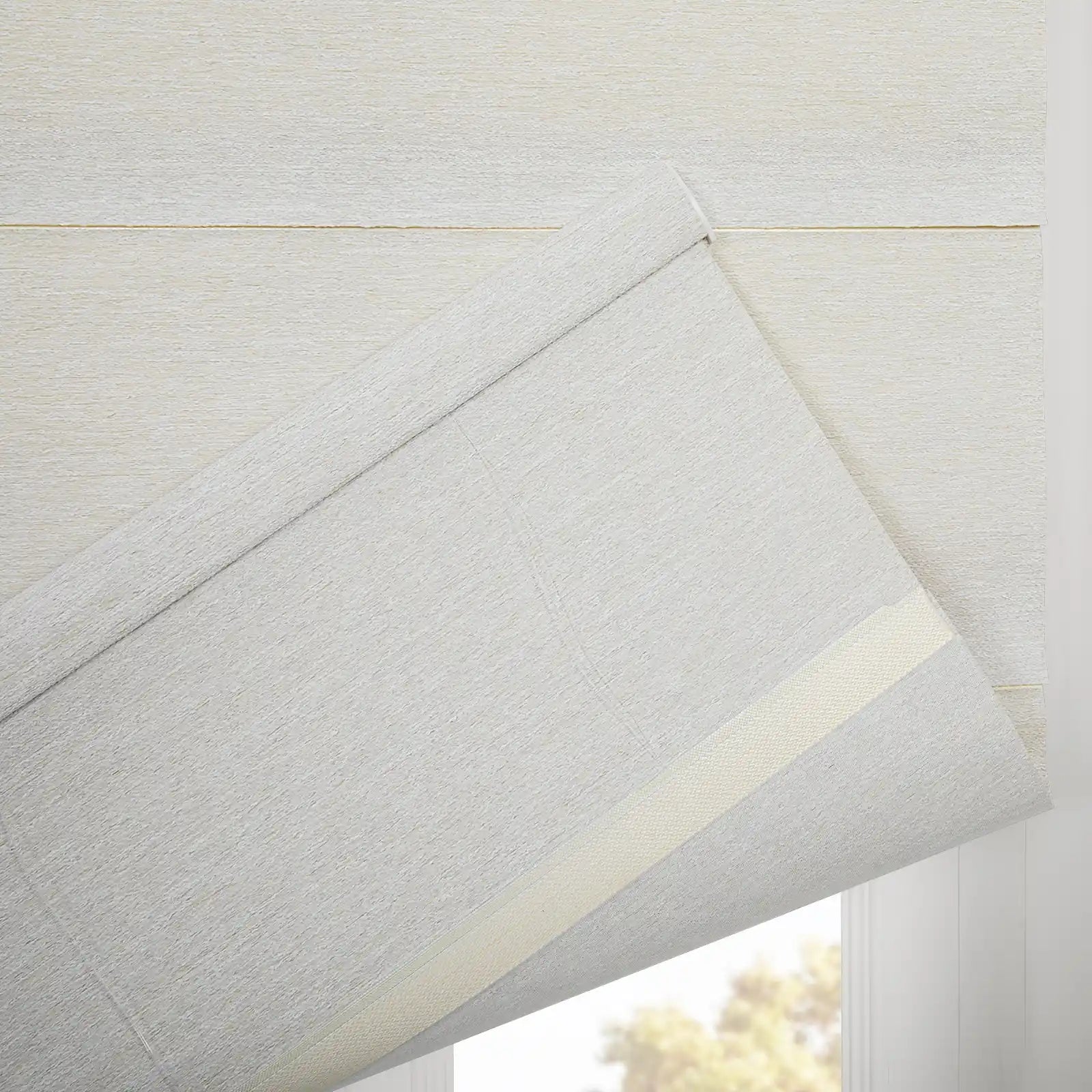 A textured light grey fabric window Blind partially unfurled, showcasing a subtle cream backing strip and a glimpse of blurred green-yellow foliage outside