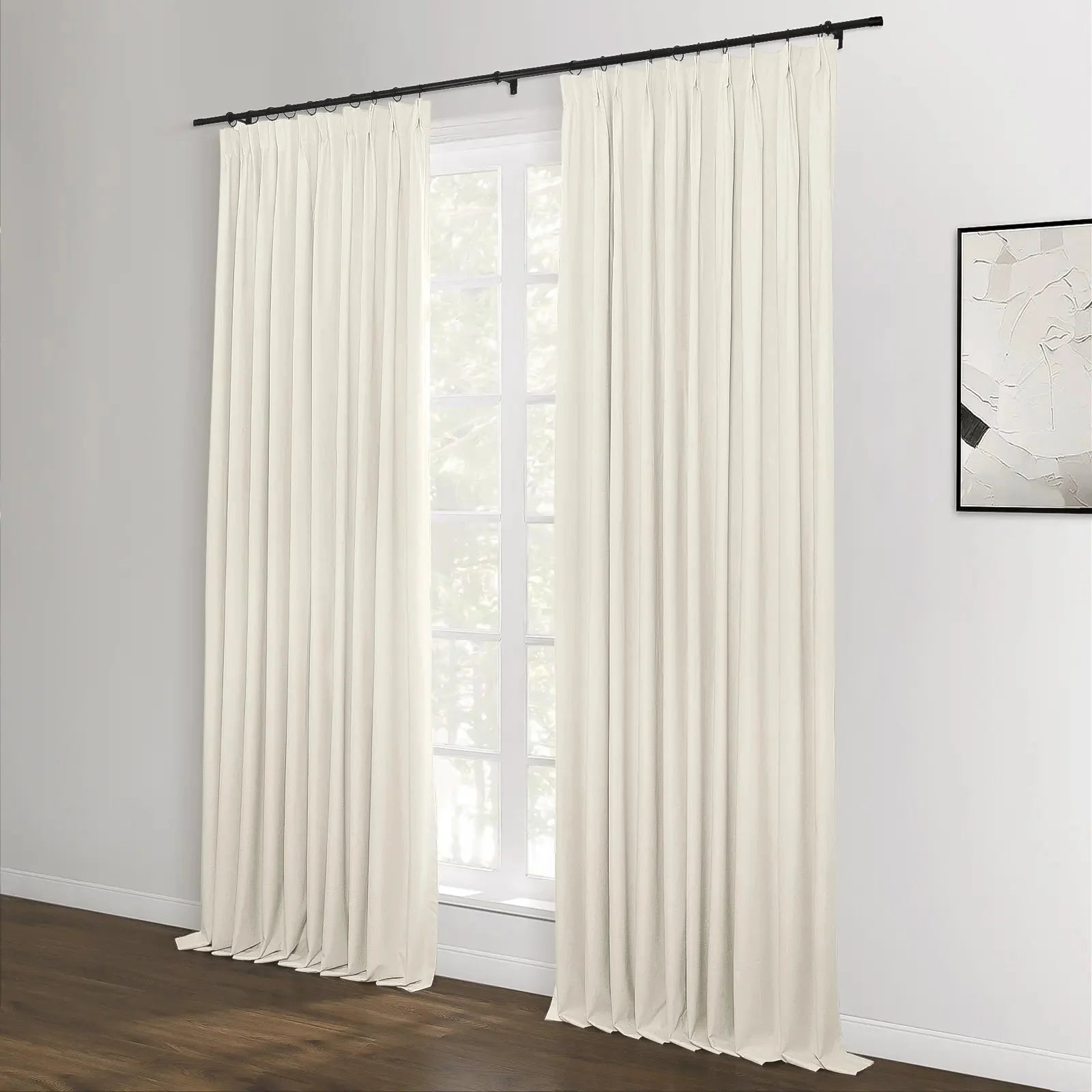 A pair of floor-length cream pinch pleat curtains on a black rod in a modern room featuring a white-paned window and a minimalist black and white abstract artwork