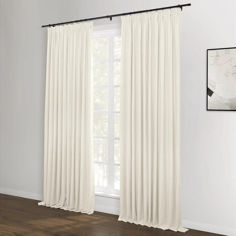 A pair of floor-length cream pinch pleat curtains on a black rod in a modern room featuring a white-paned window and a minimalist black and white abstract artwork