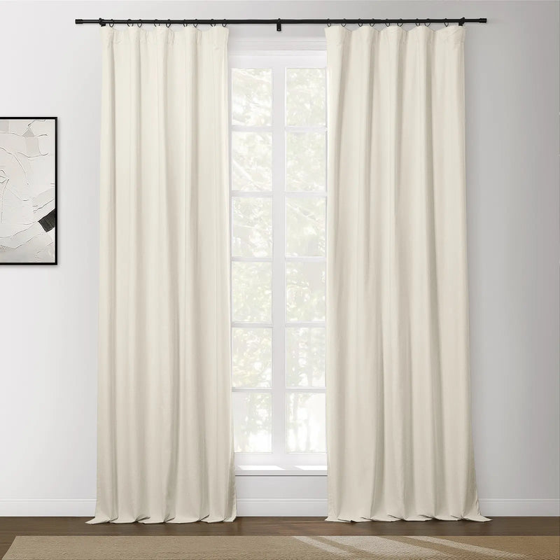 Cream blackout curtains on a black rod flank a large window overlooking trees, with a modern abstract painting on the left wall and a beige textured rug on the wood floor