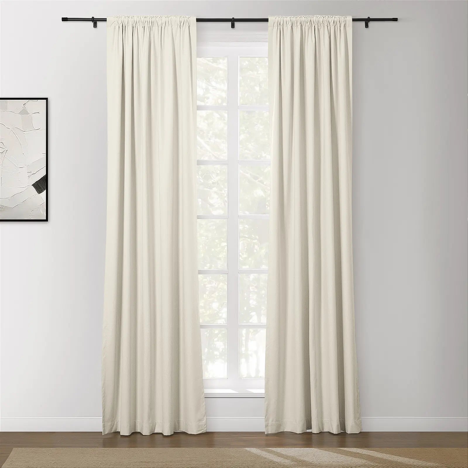 Photograph of a modern living room featuring off-white pinch-pleat drapes on a black rod framing a window with white grilles an abstract white and black artwork on the wall and a dark wood floor with a beige chevron rug