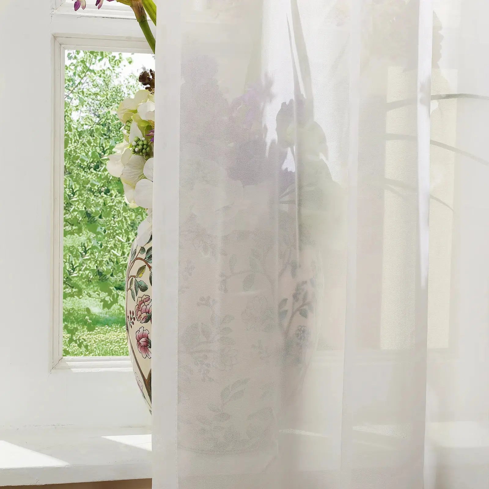 A floral patterned vase with pink and green motifs holds white and green hydrangeas on a white windowsill, partly obscured by a sheer white curtain with lush green trees visible beyond