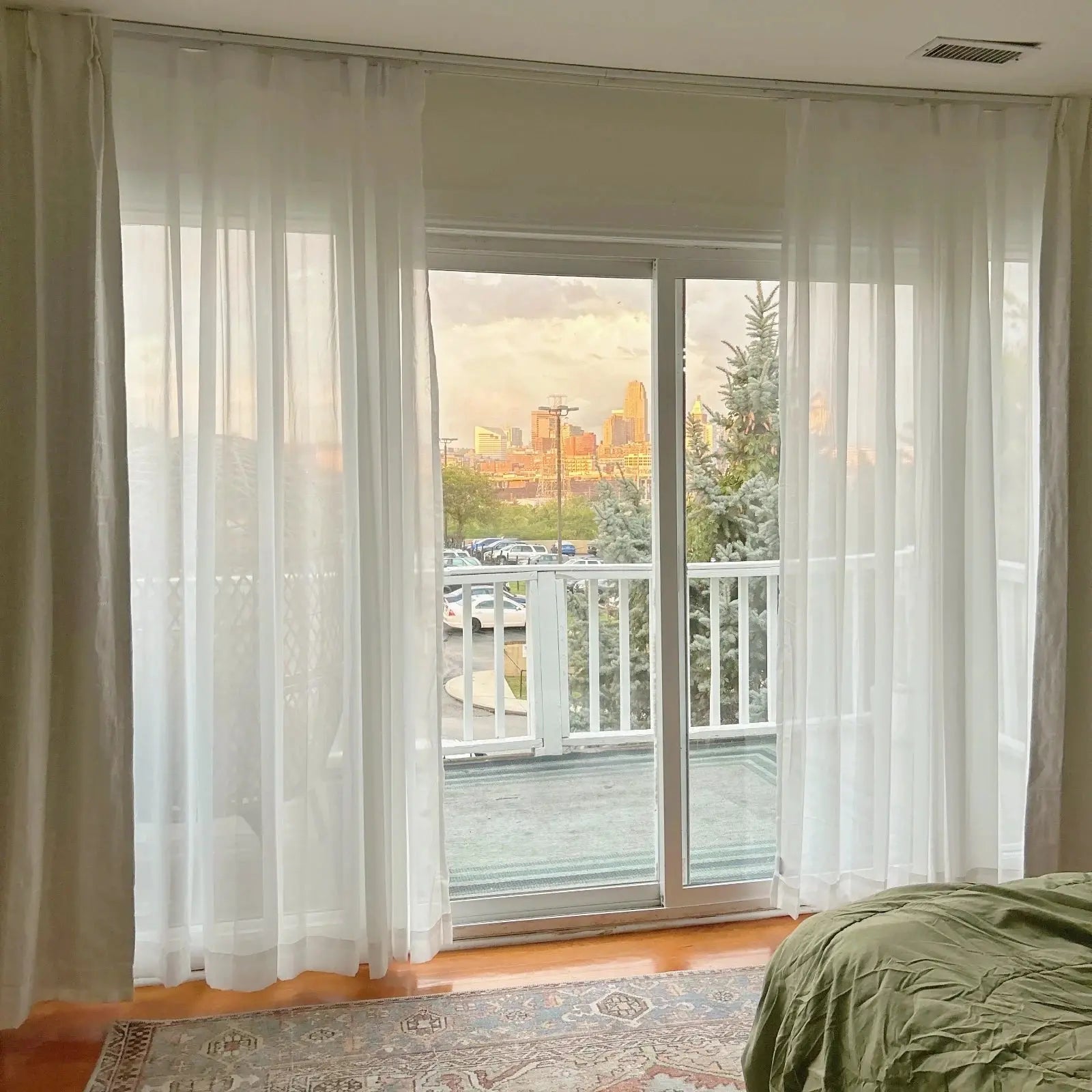 View from a bedroom window of downtown Indianapolis at golden hour, featuring the iconic One America Square and Salesforce Tower, framed by sheer white curtains, with silver-blue spruce trees and a vintage Persian rug