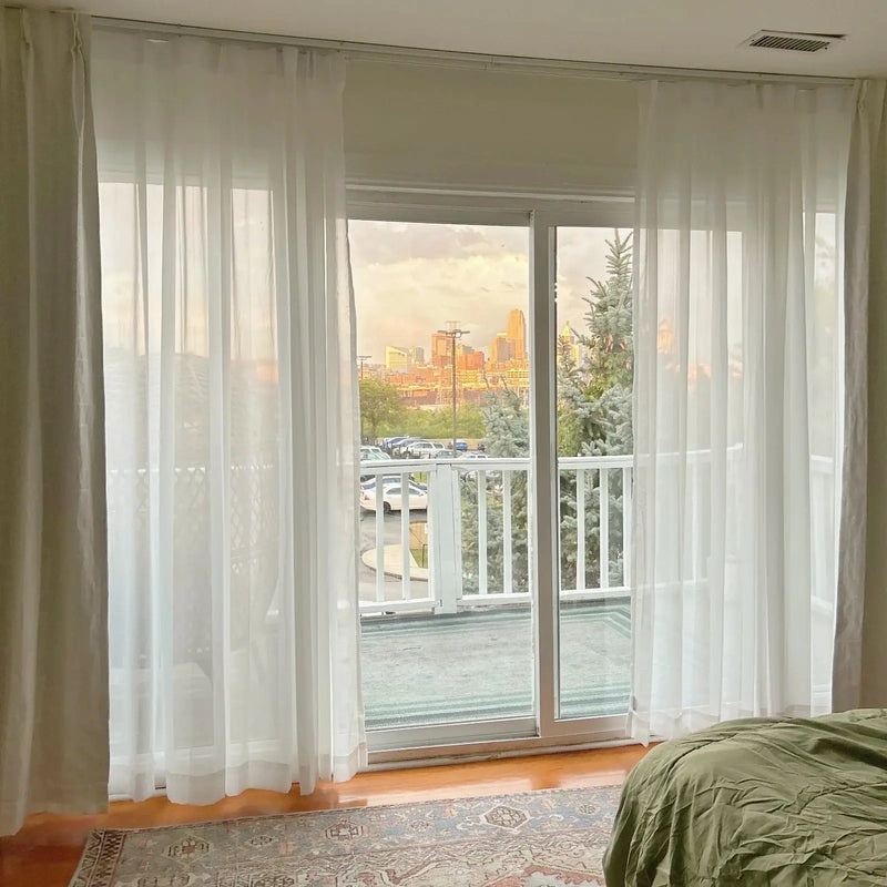 View from a bedroom window of downtown Indianapolis at golden hour, featuring the iconic One America Square and Salesforce Tower, framed by sheer white curtains, with silver-blue spruce trees and a vintage Persian rug